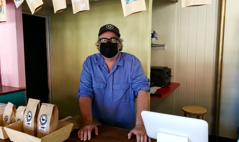 A man wearing a black face mask and blue t-shirt stands behind a cashier at a cafe.