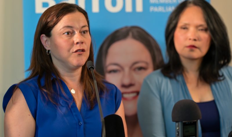 a woman with long brown hair wearing a blue blouse speaks into a microphone in front of a blue banner reading Lisa-Marie Barron with a woman in the background wearing a light blue sweater who is slightly out of focus looking on.