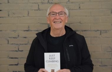 A smiling, white-haired man wearing glasses and a black jacket holds the book he authored, titled Just Keep Climbing. He stands in front of a beige brick wall.
