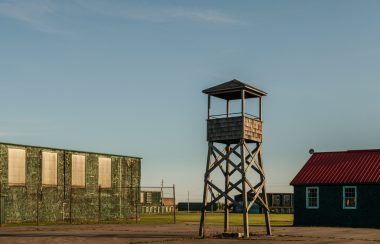 An old wooden military guard tower in the foreground with older simple barracks buildings in the background.