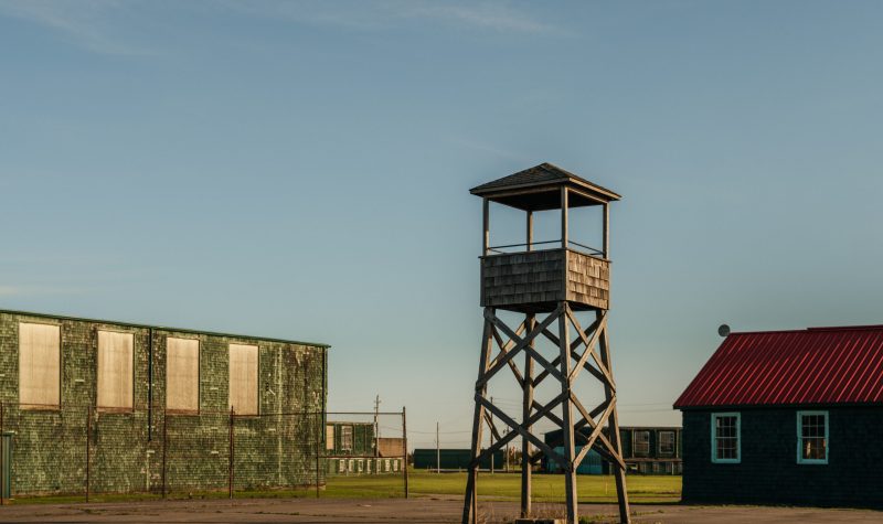 An old wooden military guard tower in the foreground with older simple barracks buildings in the background.
