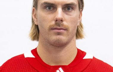 A man with blonde, medium length hair wearing a red and white hockey jersey.