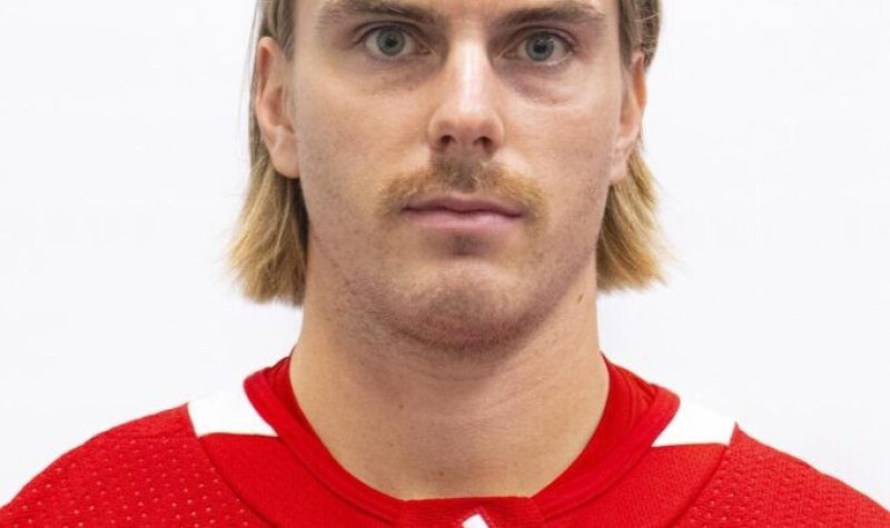 A man with blonde, medium length hair wearing a red and white hockey jersey.