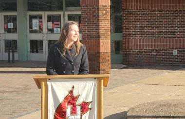 A woman stands at podium in front of a school on a sunny day.