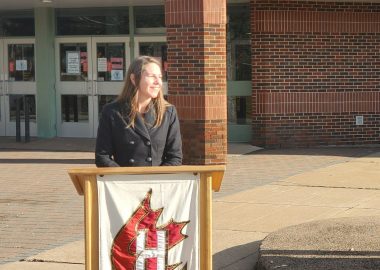 A woman stands at podium in front of a school on a sunny day.