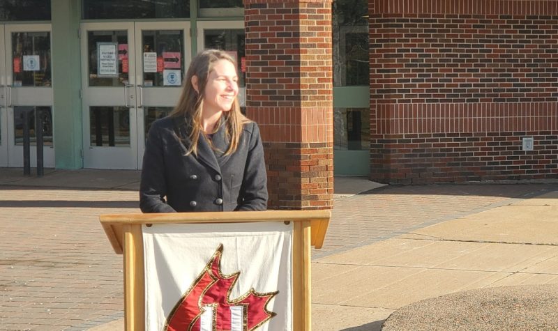 A woman stands at podium in front of a school on a sunny day.