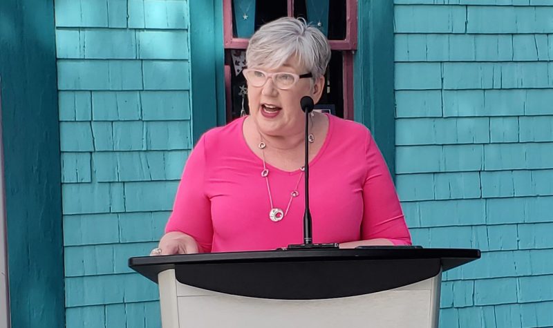 Minister Bernadette Jordan speaks at a podium in Mahone Bay against a blue house backdrop