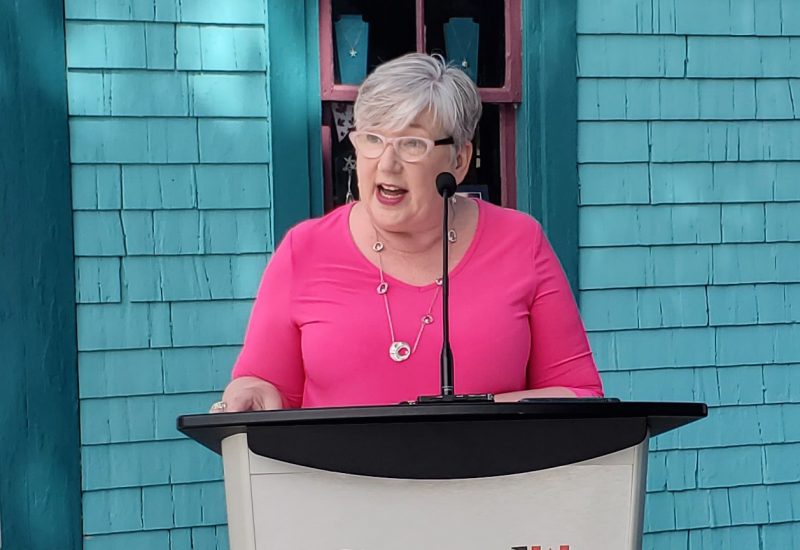 Minister Bernadette Jordan speaks at a podium in Mahone Bay against a blue house backdrop