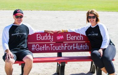 A Buddy Bench, providing people with mental health support, painted red and reading #GetinTouchforHutch seats two people on a sunny day.