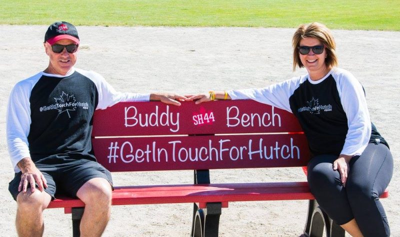 A Buddy Bench, providing people with mental health support, painted red and reading #GetinTouchforHutch seats two people on a sunny day.