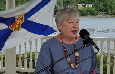 A woman speaks at podium in front of the ocean. A Nova Scotia flag flies behind her.