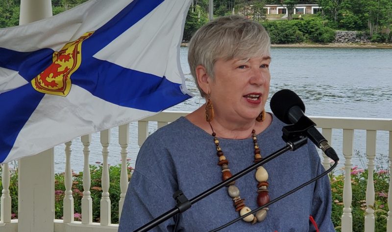 A woman speaks at podium in front of the ocean. A Nova Scotia flag flies behind her.