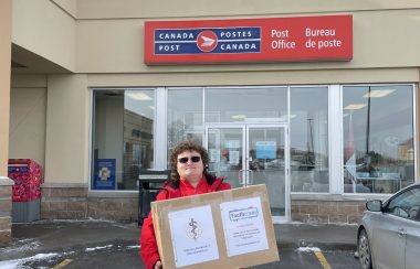 A man wearing black sunglasses and a red jacket stands in front of a Canada Post office, holding a large cardboard box.