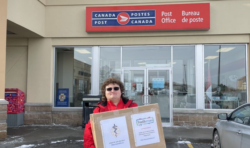 A man wearing black sunglasses and a red jacket stands in front of a Canada Post office, holding a large cardboard box.