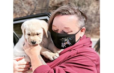 Bev Wells tenant un petit Labrador de 10 mois dans ses mains. Elle porte un masque noir avec une licorne dessus.