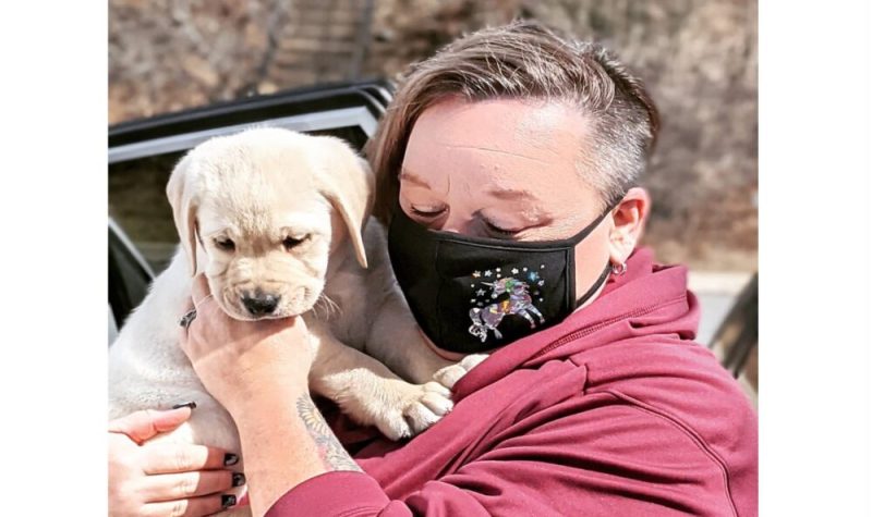 Bev Wells tenant un petit Labrador de 10 mois dans ses mains. Elle porte un masque noir avec une licorne dessus.