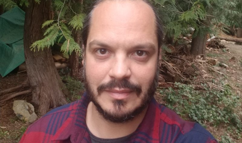 A bearded man in a red and blue plaid shirt stands in a coniferous forest.