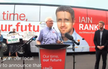 Man speaking at a red podium, with a campaign bus in the background feature Iain Rankin, leader of the Nova Scotia Liberal party. Iain Rankin stands back and to the right of the man speaking.