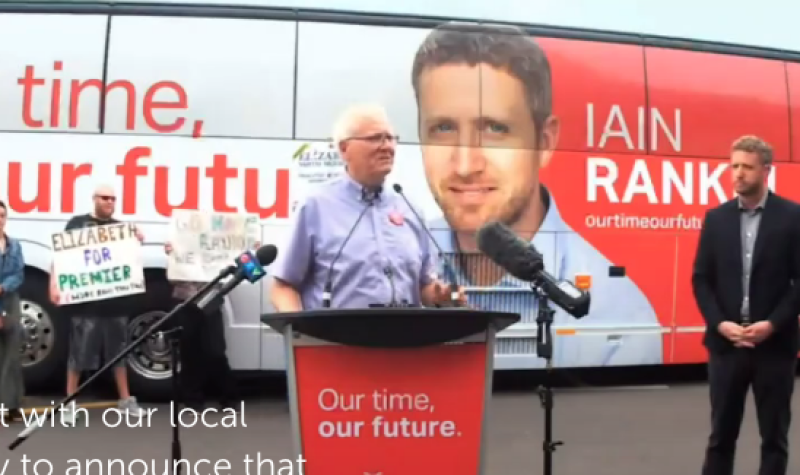 Man speaking at a red podium, with a campaign bus in the background feature Iain Rankin, leader of the Nova Scotia Liberal party. Iain Rankin stands back and to the right of the man speaking.