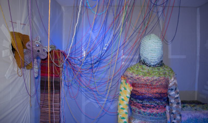A photo of Blake Hobson-Dimas in a crochet bodysuit. Threads of yarn hang from the ceiling behind him.