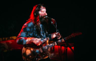 Drew Reikman stands on dark stage with a guitar.