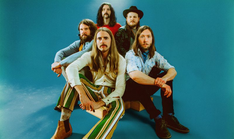 Five band members sitting with vintage looking clothes looking at the camera. There is a blue plain background behind them.