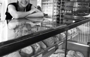 Pictured is Rousseau standing behind her bread counter. You can see a variety of breads through the class windows of the counter.