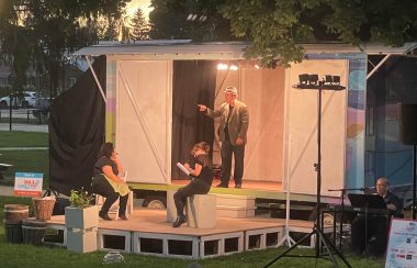 A photo at dusk showing a small stage with a smaller train boxcar like model enclosure behind it. A person with a hat on is standing inside the boxcar and pointing away from the stage, while two people are sitting on chairs in the stage in front.