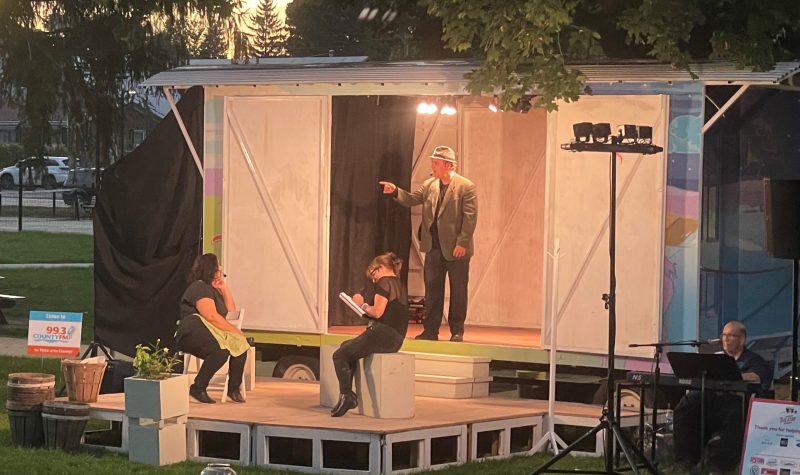 A photo at dusk showing a small stage with a smaller train boxcar like model enclosure behind it. A person with a hat on is standing inside the boxcar and pointing away from the stage, while two people are sitting on chairs in the stage in front.