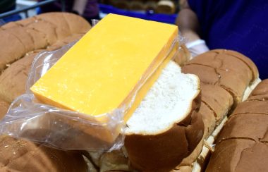 Large pieces of white bread with brown crust and a block of orange cheddar wrapped in plastic sits on top of a piece of thick sliced bread