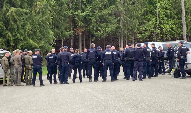 Around 31 RCMP in blue and army green uniform congregate in a forest clearing.