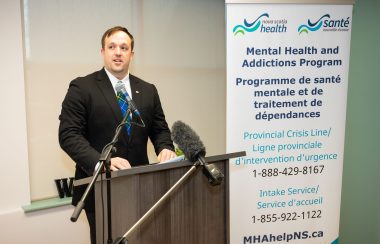 Brian Comer, the Minister responsible for the Office of Addictions and Mental Health. He is seen standing behind the podium with a microphone. There is a mental health addiction program poster behind him.