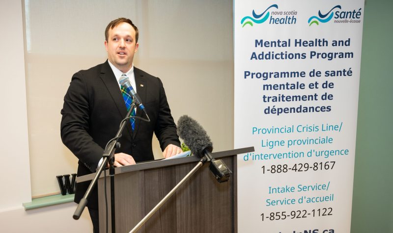 Brian Comer, the Minister responsible for the Office of Addictions and Mental Health. He is seen standing behind the podium with a microphone. There is a mental health addiction program poster behind him.