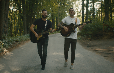 Two young men playing acoustic guitars while walking down the middle of a wooded road.