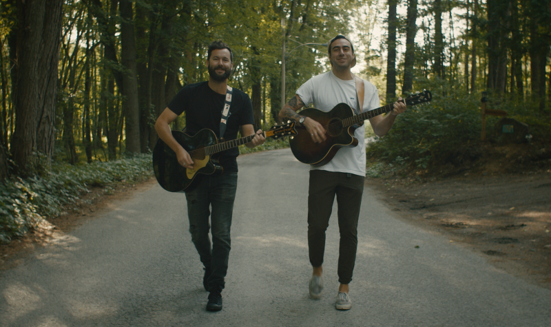 Two young men playing acoustic guitars while walking down the middle of a wooded road.
