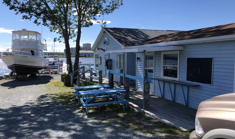 Exterior of white marina building. In the background, there is a large boat and a shoreline and beach.