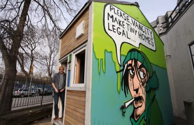 Bryn Davidson stands outside of a wooden tiny home with an artwork painted on the side featuring a man smoking against a green background
