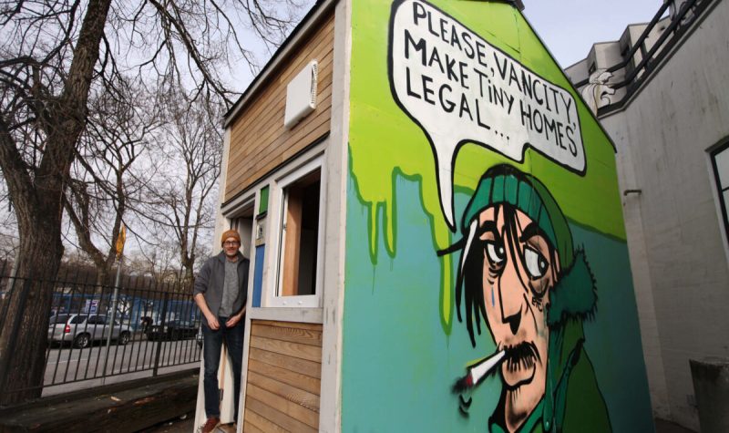 Bryn Davidson stands outside of a wooden tiny home with an artwork painted on the side featuring a man smoking against a green background