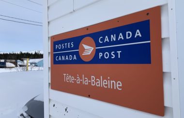 Un panneau aux couleurs de Postes Canada indiquant Tête-à-la-Baleine est accroché sur un mur extérieur.