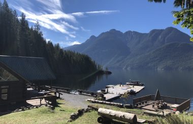 Sun beams down on lawn, deck and dock beside a waterfront building with mountains in the background.