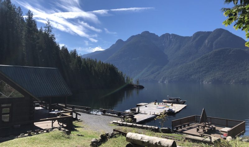 Sun beams down on lawn, deck and dock beside a waterfront building with mountains in the background.