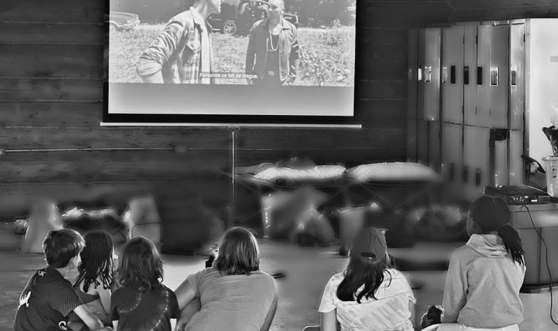 Six enfants assis devant un encrant projecteur, regardant une image ou il y a deux hommes.