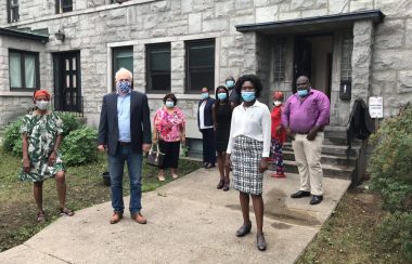 Denis Trudel avec des membres de la communauté noire davant un bâtiment en pierre grise. Les gens portent tous masqués.