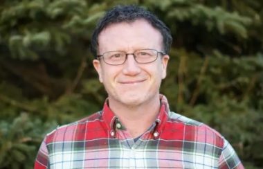 A man wearing a red, white and black flannel shirt with glasses and short-cropped hair smiles in front of a background of foliage.