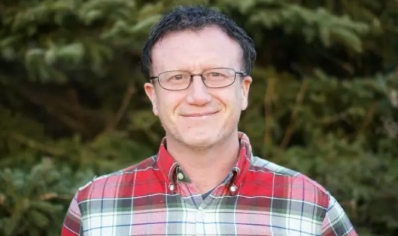 A man wearing a red, white and black flannel shirt with glasses and short-cropped hair smiles in front of a background of foliage.