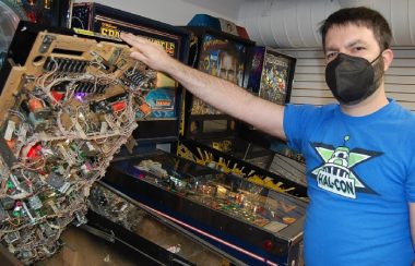 A man in a blue T-shirt wearing a black face-mask holds up the playfield or surface of a pinball machine, revealing a network of electronics.