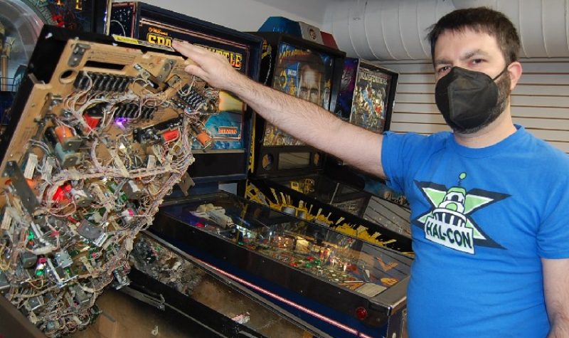 A man in a blue T-shirt wearing a black face-mask holds up the playfield or surface of a pinball machine, revealing a network of electronics.