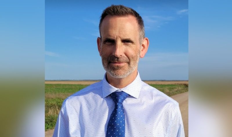 A man with close-cropped hair and a light beard wearing a white button-up shirt and a blue necktie is pictured against a blue sky and what appears to be a field on a farm on a country road.