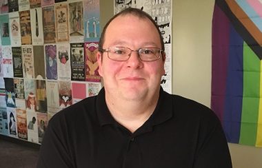 A man with glasses and a black V-neck shirt and short hair is pictured in the poster-covered hallway of CHMA FM in Sackville, with a rainbow flag on the wall.