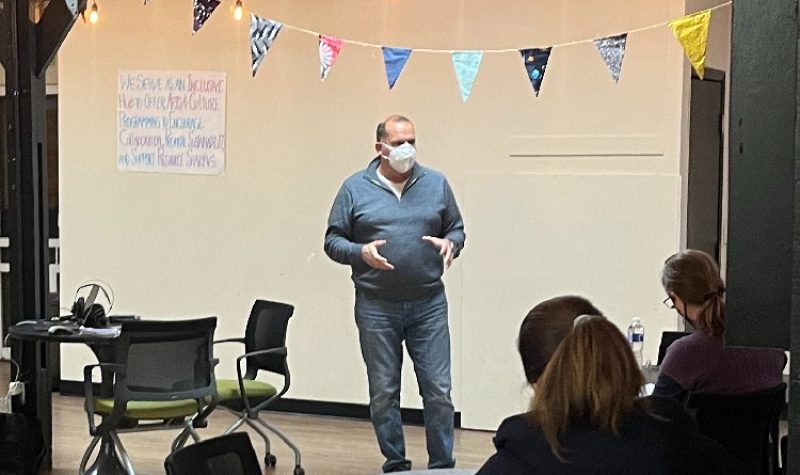 A man wearing a face mask gestures as he speaks in front of a group of people who are seated in a room.